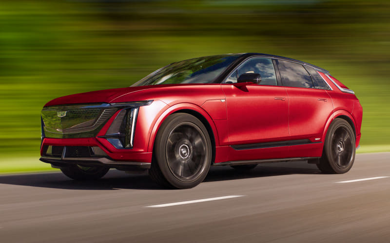 Red Cadillac SUV driving at speed on a two-lane road with a green blur background, front grille and emblem visible in motion.