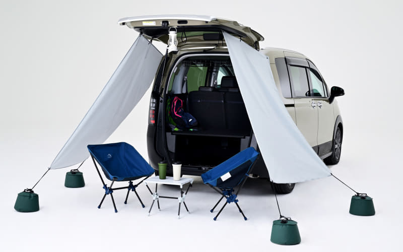 Camper van with rear hatch open and a light canopy tent anchored to the ground, plus blue chairs and a small table set up outside.