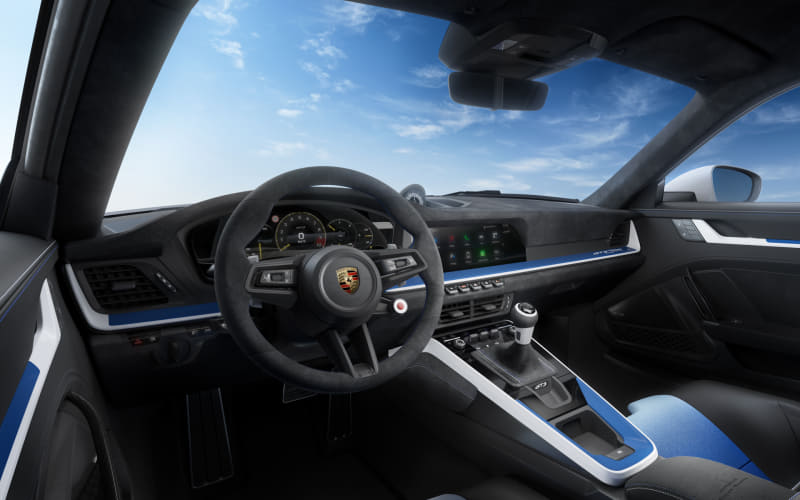 Driver’s-seat view of a Porsche interior with a multi‑function steering wheel, digital gauges, and blue accent trim.