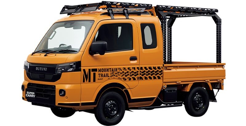 Orange Suzuki Super Carry pickup with roof racks and a black ladder frame, side decals reading 'MT Mountain Trail' in black, viewed at an angle for a rugged work vehicle context.