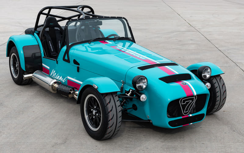 Teal open-top sports car with pink racing stripe, parked on a concrete surface on a sunny day. Recounted details: two seats and roll bar visible in the cabin.