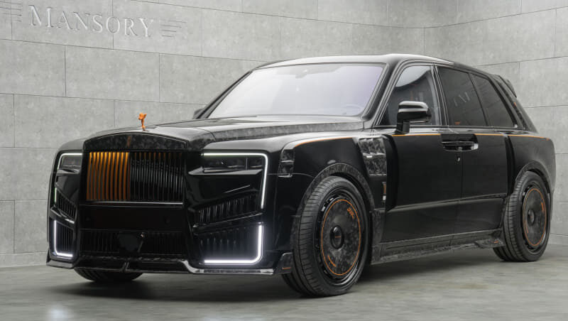 Black luxury SUV with orange accent trim, customized by Mansory, parked in a showroom with a gray stone wall in the background