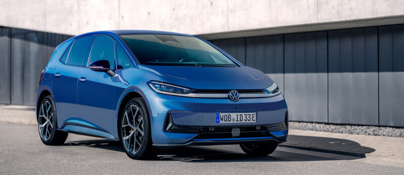 Front three-quarter view of a blue Volkswagen ID.3 electric hatchback parked on a paved area beside a modern concrete building