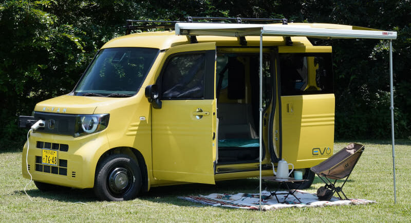 Yellow camper van with open side door and awning set up on grass, a rug and a chair laid out for camping.