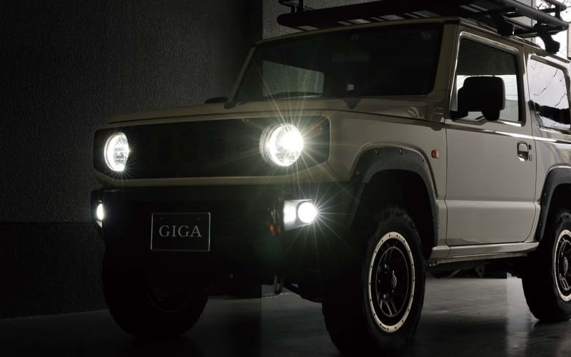 Front view of a white off-road SUV with bright headlights on in a dark garage, roof rack visible and 'GIGA' license plate.
