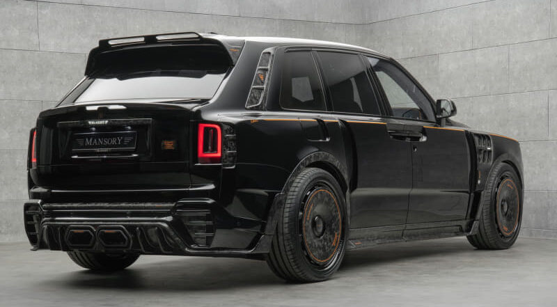 Black Mansory-modified SUV in a showroom, rear three-quarter view with tinted windows and orange-accent wheels, glossy finish.