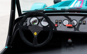 Close-up of a race car cockpit showing a black steering wheel, gauges, and dashboard controls with blue exterior visible through the window.