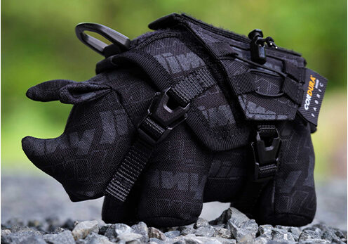 Small black dog wearing a tactical harness with straps on a rocky path, with a blurred green background