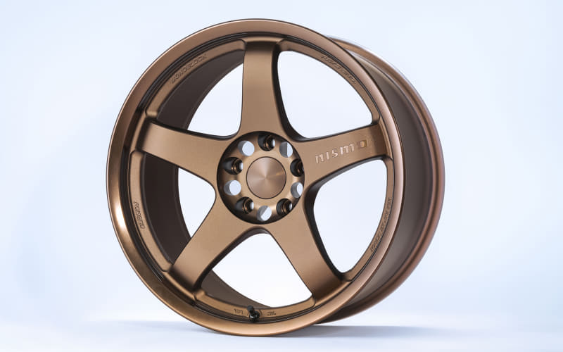 Bronze alloy car wheel with five spokes photographed against a light blue background, showing the bolt hub in the center and a Nismo logo on the spoke.