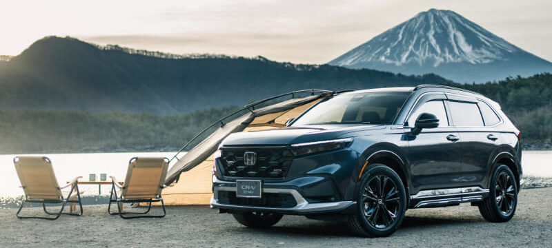 Blue Honda CR-V SUV parked on gravel at a lakeside campsite with chairs and a tent, mountain in the background.