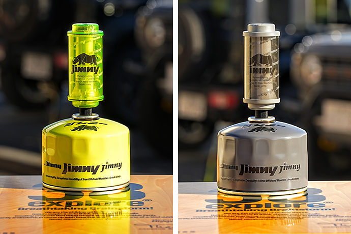 Two small camping fuel canisters on a wooden table, left yellow with a matching green top and the word 'jimny' on the body, right a silver/gray version with a similar label; blurred vehicles in the background.