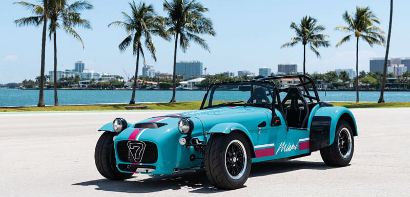 Turquoise vintage open-top sports car with a pink stripe parked by a palm-lined waterfront.