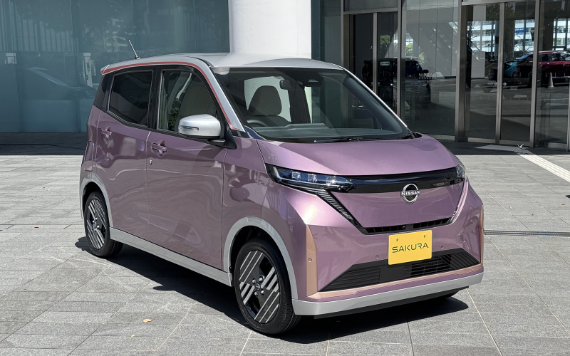 Lavender Nissan minivan parked outside a dealership, front three-quarter view with chrome accents and a yellow Sakura license plate.