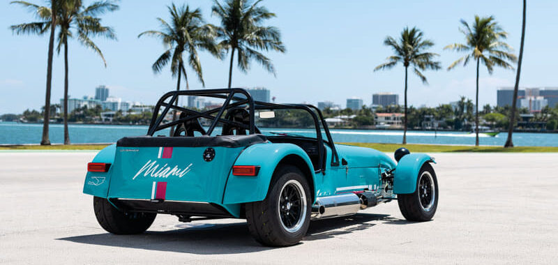 Turquoise vintage roadster with open cockpit parked on a sunlit coastal road; palm trees line the water in the background with a distant city skyline.