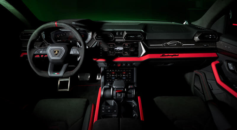Inside a Lamborghini sports car: steering wheel, digital gauges, central touchscreen, and red ambient lighting along the dashboard and center console.
