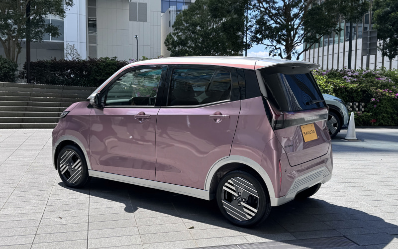 Pink compact hatchback seen from the rear side, parked on light paving near a building, with a white roof and dark-tinted windows (Sakura model).