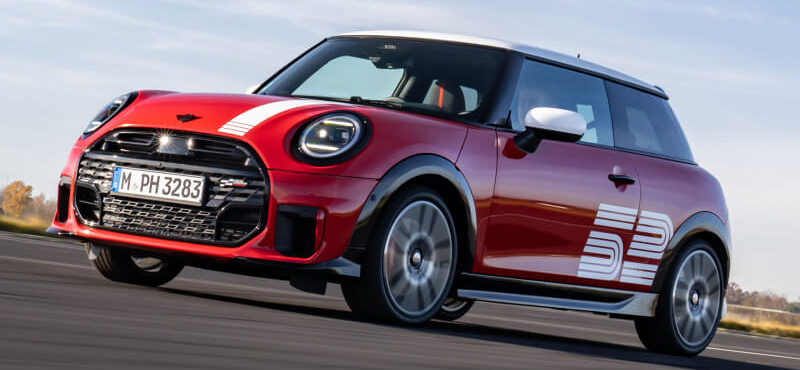 Red Mini Cooper in motion on an open road, white roof and side graphic, blue sky above, motion blur behind the wheels