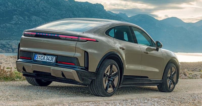 Beige Porsche Taycan parked on a gravel overlook with mountain range and lake in the background.