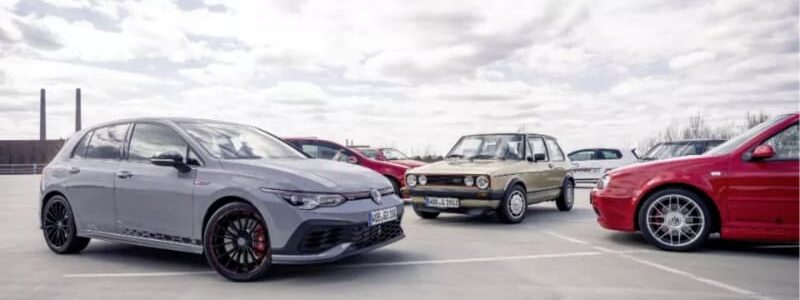 Grey modern hatchback parked in a lot, with a vintage beige car and a red sports car nearby under a cloudy sky