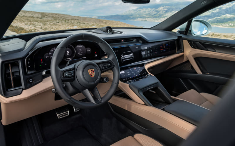 Interior of a Porsche with tan leather seats, digital instrument cluster, and a large center touchscreen. Outside view shows a scenic landscape through the windows.