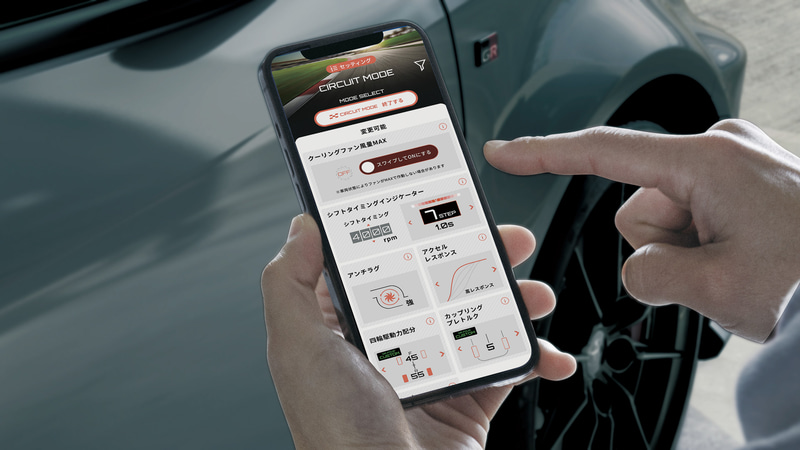 006_l - 自社ローン中古車販売専門店 ファンカーズ Person holds a smartphone displaying a car tuning app in Circuit Mode beside a car door panel and wheel.