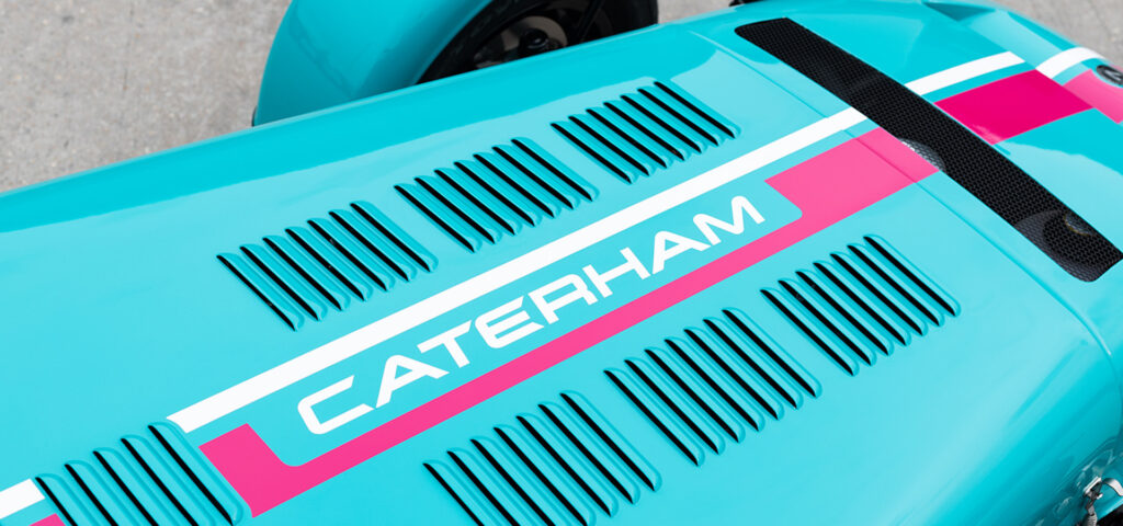 Close-up of a turquoise vintage race car's rear deck with black vent slits and bold pink stripe accents on a gray pavement background.