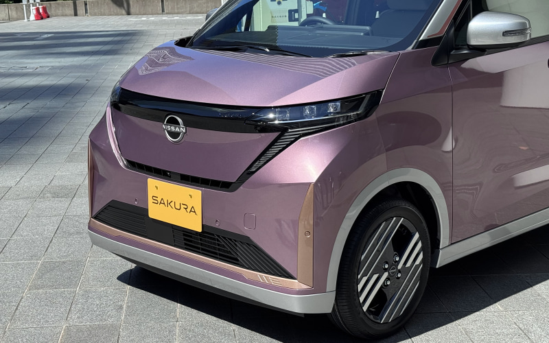 Front-left view of a pink Nissan Sakura electric car with a yellow license plate in a parking lot; visible chrome grille and alloy wheel.
