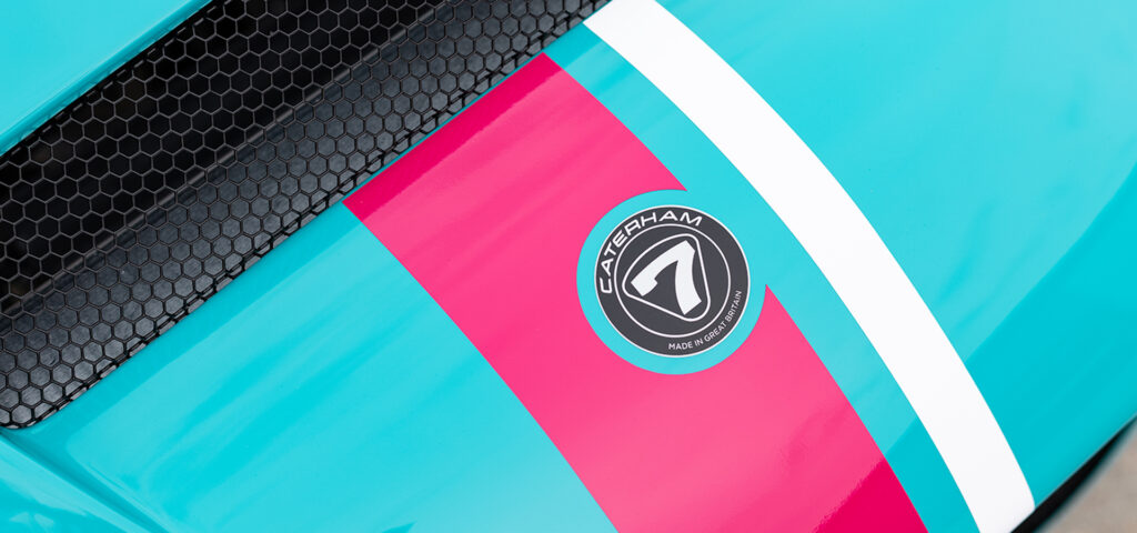 Close-up of a turquoise car hood with a pink racing stripe and black honeycomb grille, featuring a circular Caterham badge in the stripe area.