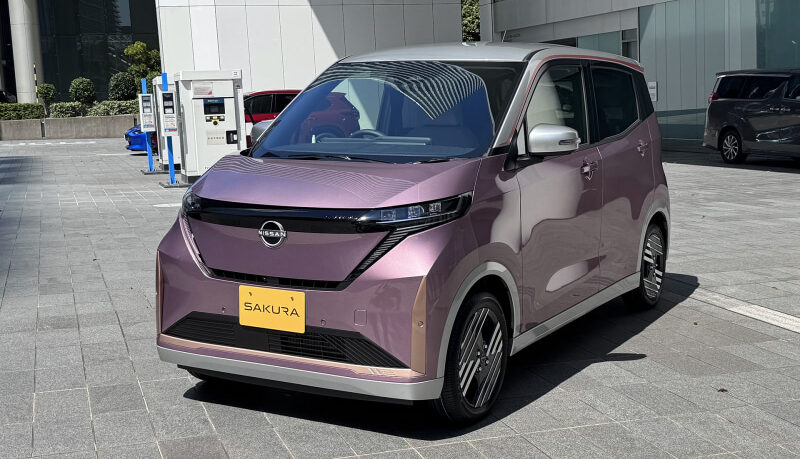 Front three-quarters view of a pinkish-purple Nissan Sakura hatchback parked on a paved plaza near a modern building. The car has a yellow license plate labeled 'SAKURA' and black grille with Nissan emblem.