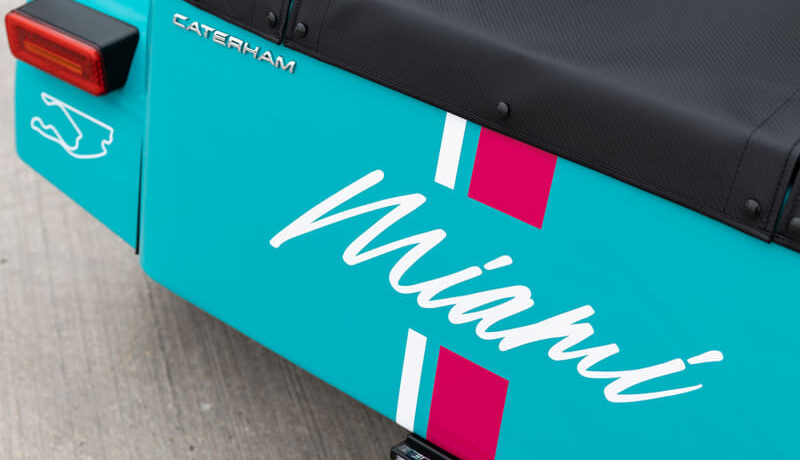 Close-up of a turquoise Caterham vehicle rear with a white cursive logo and pink accent stripes.