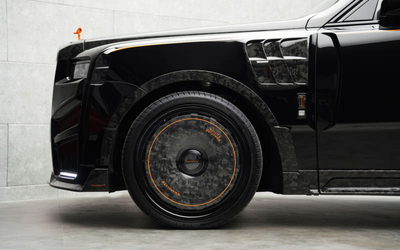 Close-up of a black car's front left wheel with a carbon-fiber style rim and orange accent lines on a light gray garage wall backdrop, highlighting the tire and fender area.