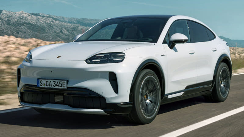 White Porsche electric SUV driving on a highway with mountains in the background (three-quarter front view).