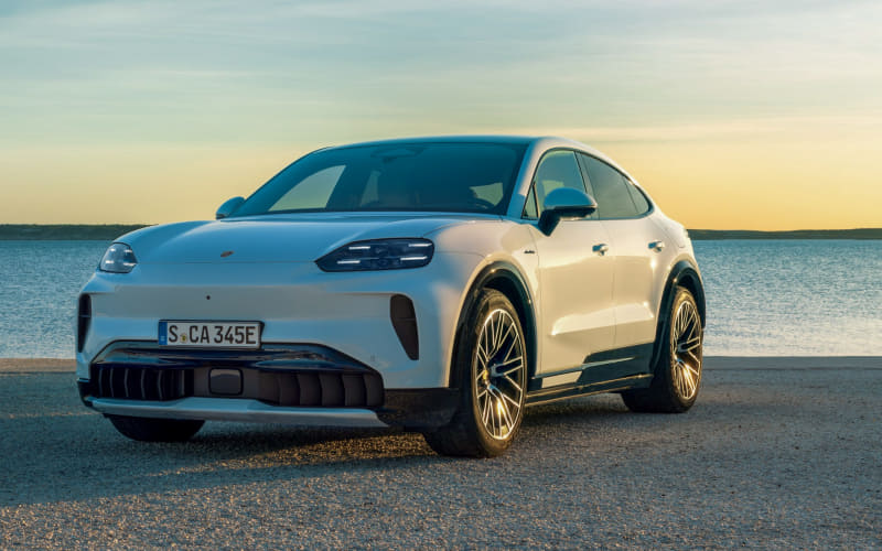 White Porsche SUV parked on a coastal road at sunset with calm water in the background.
