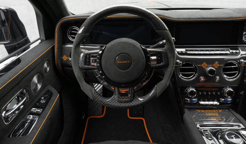 Luxurious car interior with a carbon fiber steering wheel, orange stitching, and touchscreen dashboard behind it.