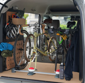 Two road bikes secured in the back of a vehicle using a bike rack, with helmets and water bottles nearby in the cargo area.