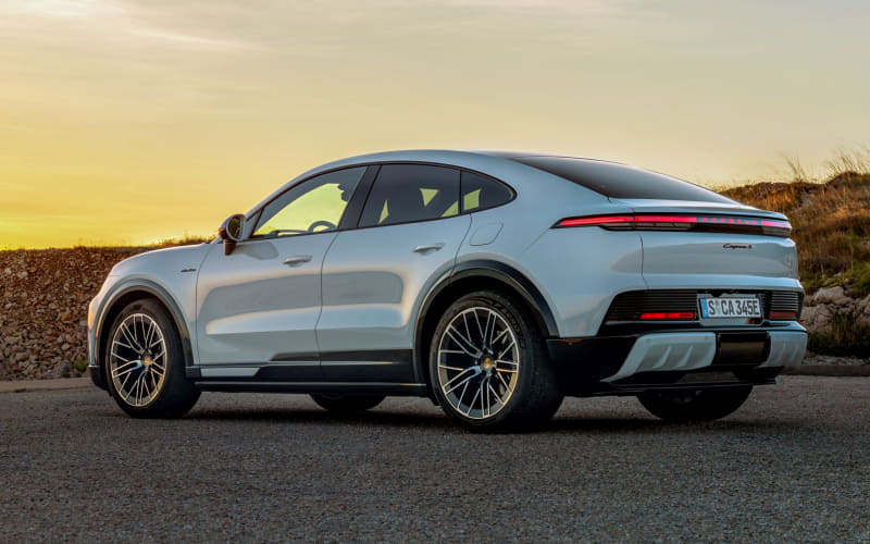 Rear three-quarter view of a silver Porsche Cayenne parked on a paved road at sunset, with a low stone wall in the background.