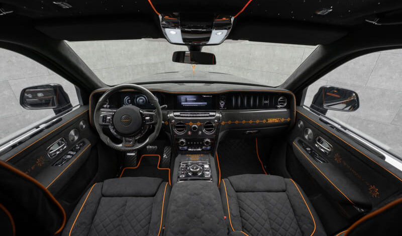 Front interior of a luxury car with black quilted seats and orange piping, featuring a digital dash and center console visible from the driver’s seat route. Optional descriptive: wide shot shows steering wheel and touchscreen.
