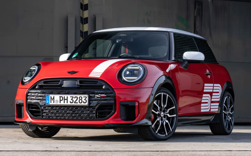 Front three-quarter view of a red Mini Cooper with a white roof and black racing stripes on the hood, parked outdoors.