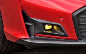 Close-up of the front left corner of a red car, showing a yellow fog light embedded in the lower bumper.