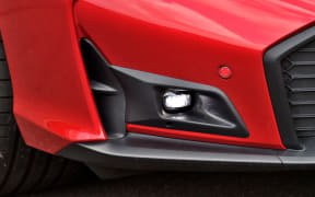 Close-up of a red car's front bumper showing a recessed fog light and black trim surrounding it, with part of the tire visible on the left.