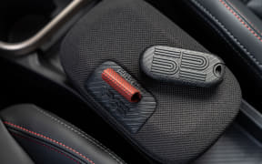 Two key fobs resting on a textured car seat pocket. One is red and cylindrical, the other is a black, ribbed fob with a button pattern.