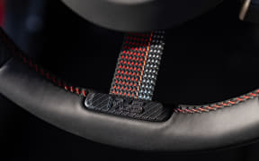 Close-up of a car steering wheel with a woven red-and-gray grip and black leather stitching.Se