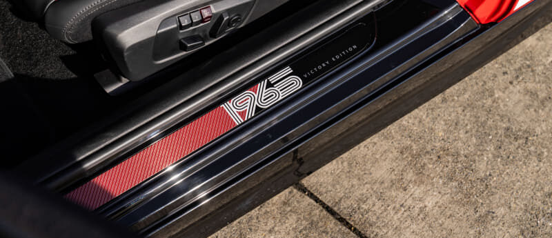Car door sill with a '1965 Victory Edition' badge, red interior accents, and seat stitching visible nearby.