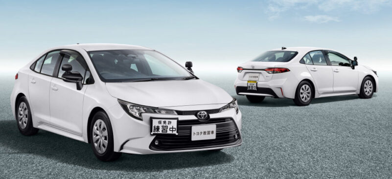 Two white Toyota sedans on a wide open lot with a clear blue sky; the front car has practice plates, with another car in the background.