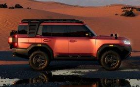 Orange off-road SUV with roof rack parked on a desert dune at sunset, reflecting on wet sand nearby.