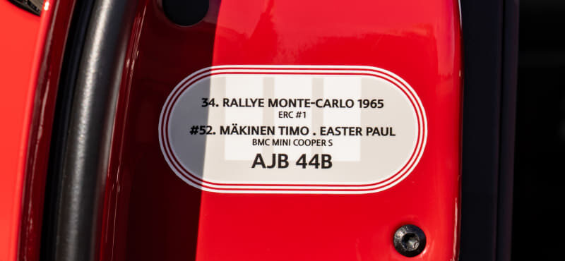 Close-up of a red car’s body with a white oval label listing rally details: “34. Rallye Monte-Carlo 1965 ERC #1” and “#52. Mäkinen Timo . Easter Paul BMC Mini Cooper S AJB 44B.”
