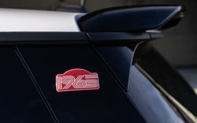 Rear quarter panel of a dark blue car with a pink '1965' badge and a spoiler above.