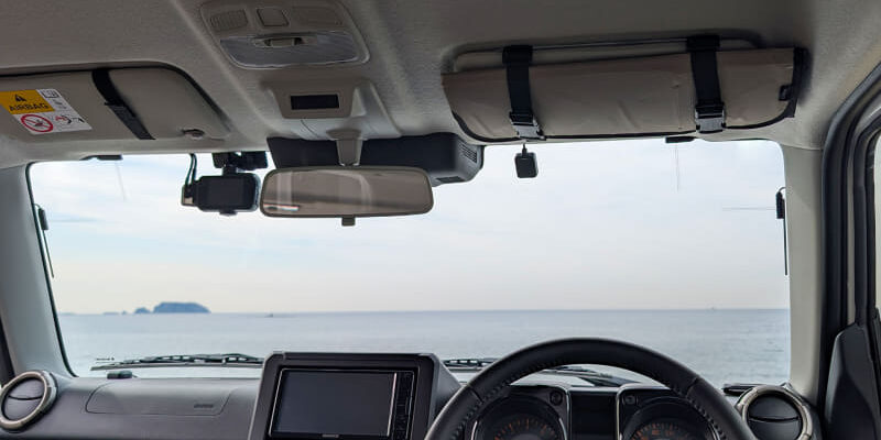 Interior view of a car cabin with the steering wheel and dashboard, looking out at a calm sea and distant island through the windshield.