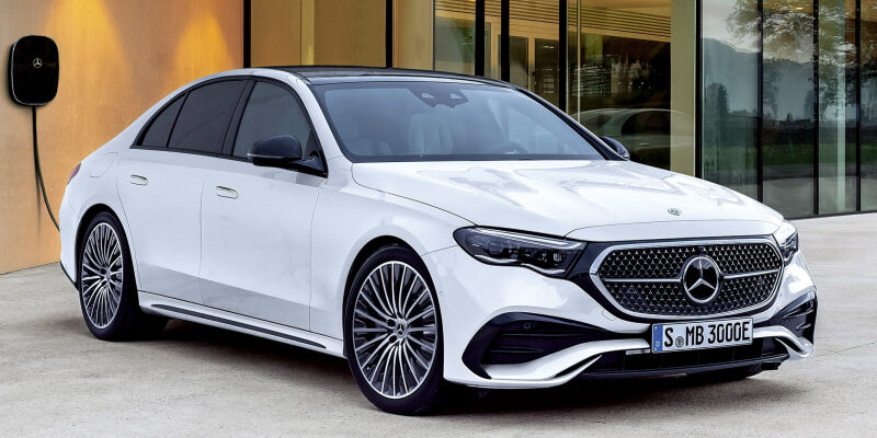 White Mercedes-Benz sedan parked in front of a glass-walled building, featuring a diamond grille and large multi-spoke wheels.