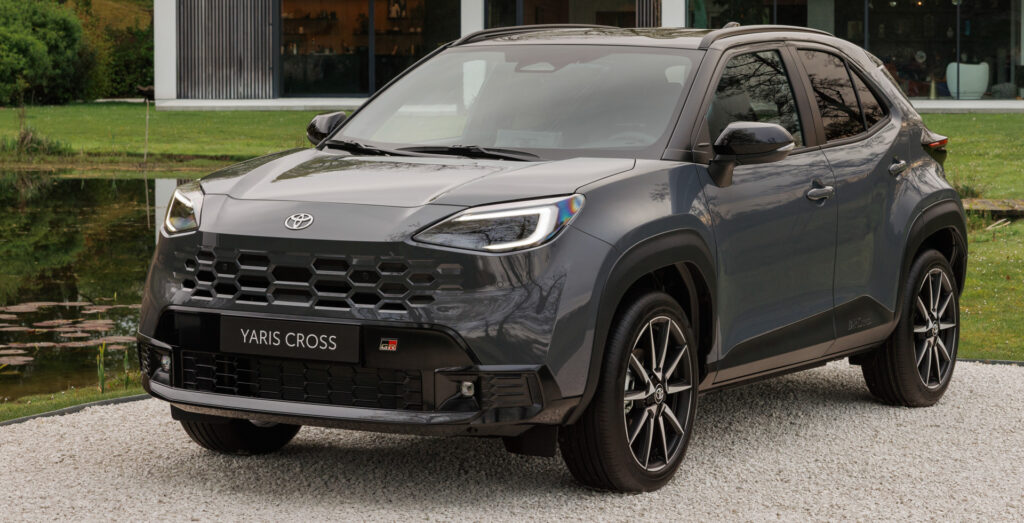Front-left view of a gray Toyota Yaris Cross SUV parked on a gravel path with modern glass building in the background.